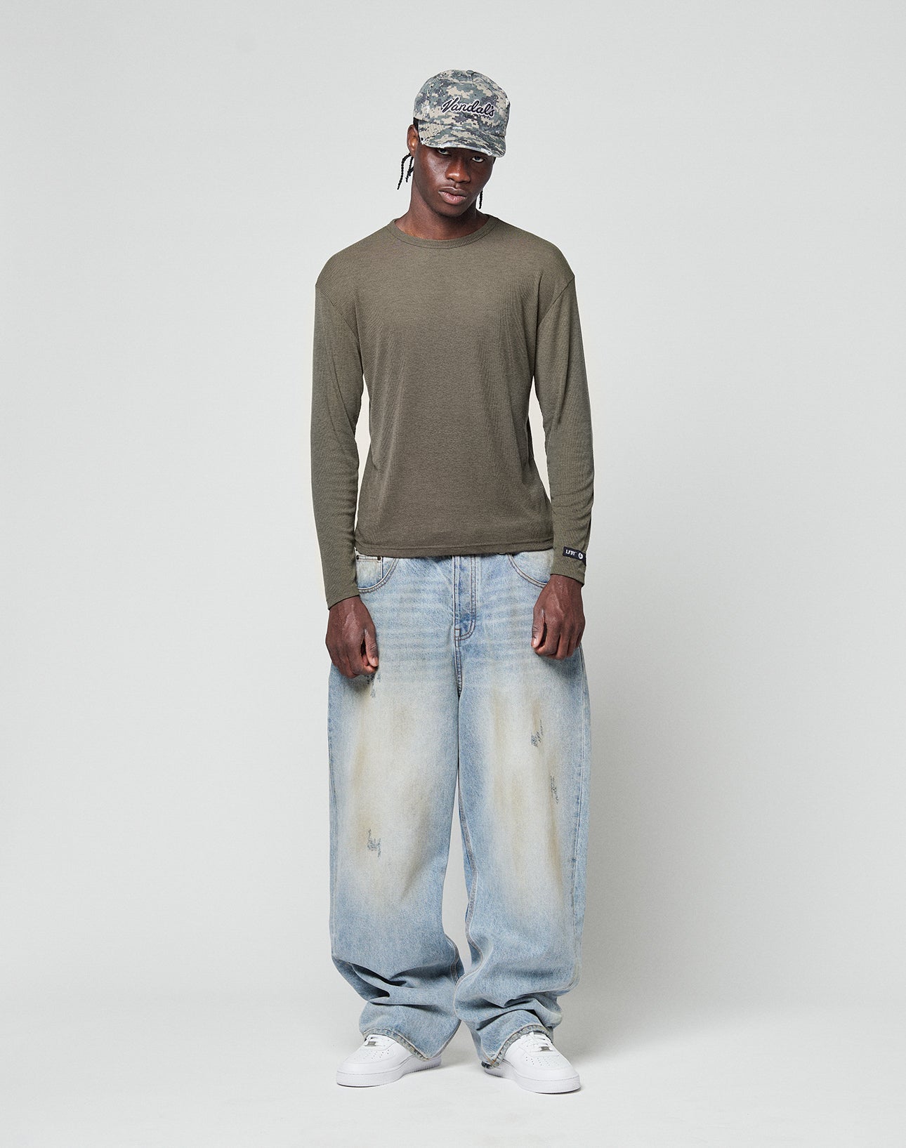A person wearing the LIVE FAST DIE YOUNG LFDY Rib Longsleeve in olive green, loose light blue jeans, white sneakers, and a blue patterned cap stands against a plain light gray background.
