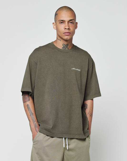 A person with a shaved head and tattoos stands against a plain background, wearing the LIVE FAST DIE YOUNG Basic +49 Chest Tee—an oversized olive green 100% Baumwolle shirt with a subtle logo—paired with light shorts, hands in pockets.