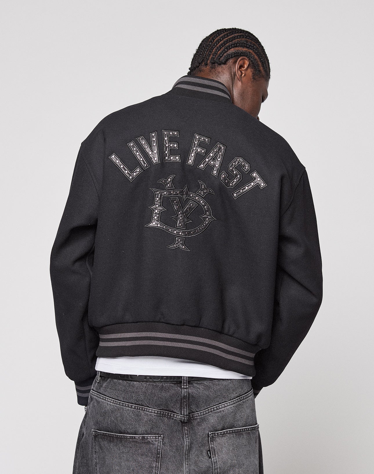 A person with braided hair faces away, wearing the LIVE FAST DIE YOUNG Live Fast College Jacket featuring a Nieten Patch Logo and LIVE FAST emblem on the back, striped cuffs and collar, metal zip, paired with faded black jeans.