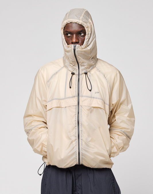 A person wearing the LIVE FAST DIE YOUNG Live Fast Nylon Jacket in light beige with reflective print, hood up and hands in pockets, stands against a plain light gray background.