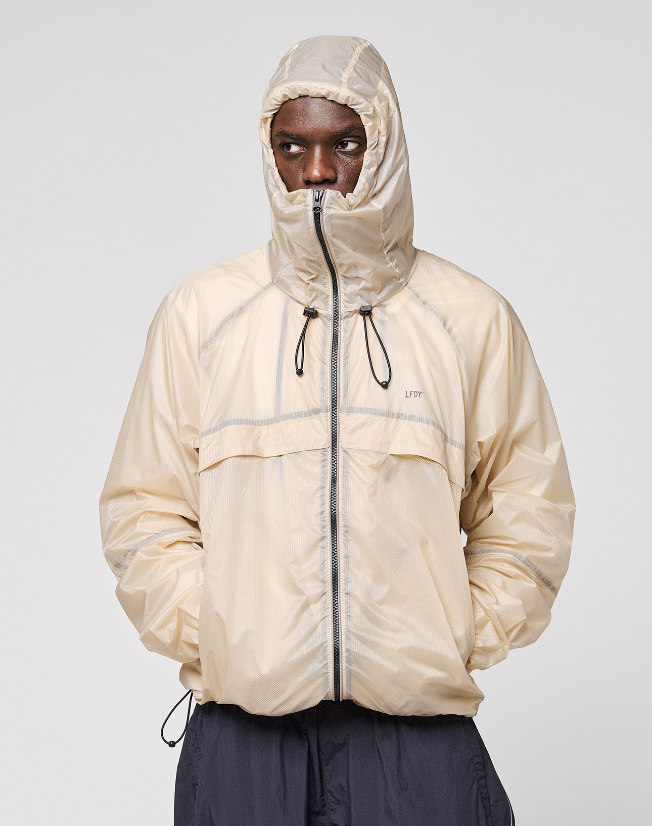 A person wearing the LIVE FAST DIE YOUNG Live Fast Nylon Jacket in light beige with reflective print, hood up and hands in pockets, stands against a plain light gray background.
