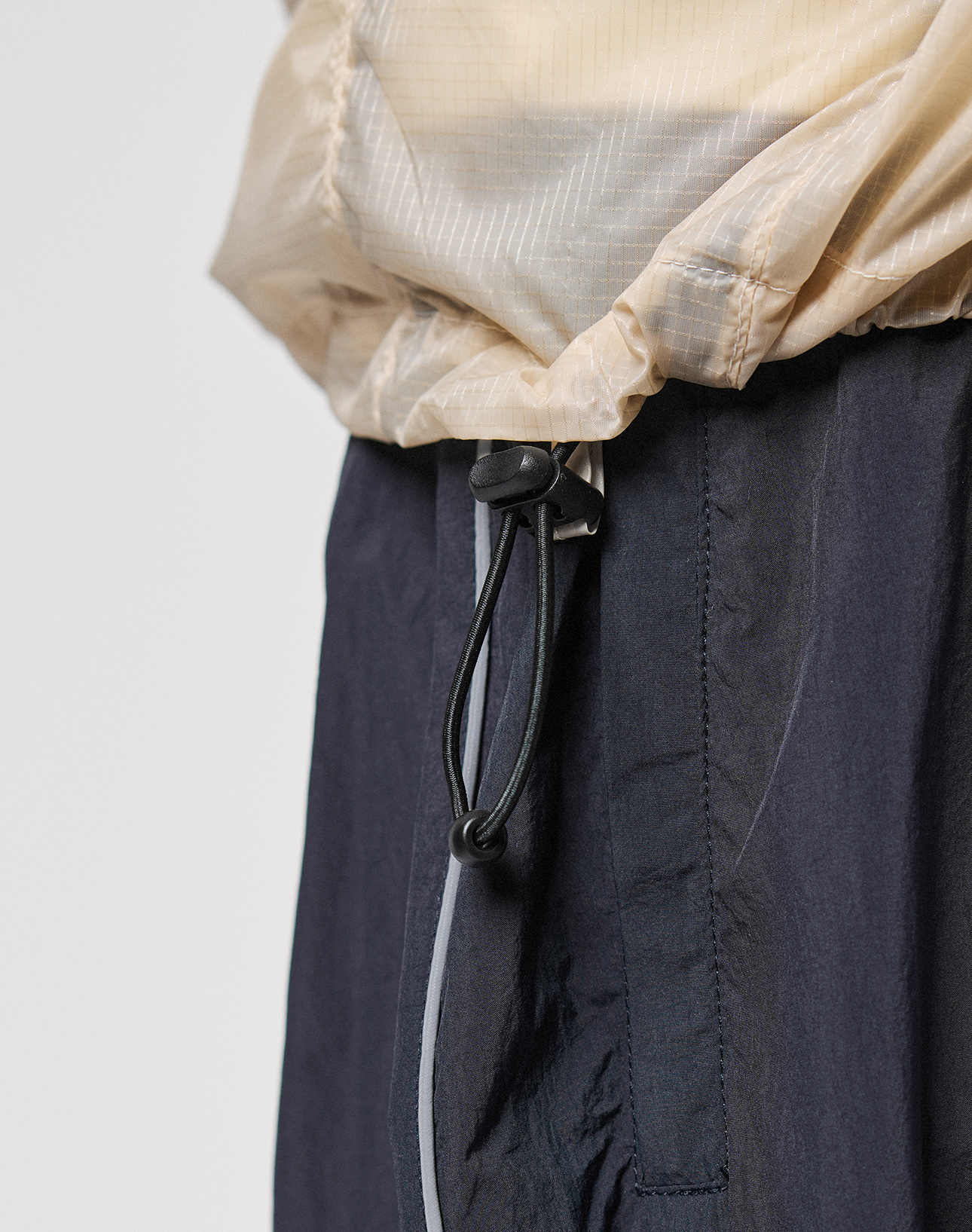 Close-up of a person wearing the Live Fast Nylon Jacket by LIVE FAST DIE YOUNG in light beige with a black drawstring, paired with dark pants featuring seam and pocket details.