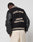 A person stands against a light background wearing cream pants and the LFDY x Schott NYC Varsity Jacket by LIVE FAST DIE YOUNG, featuring beige LIVE FAST and LÉQUIPE CÔTE DAZUR lettering on the black back.