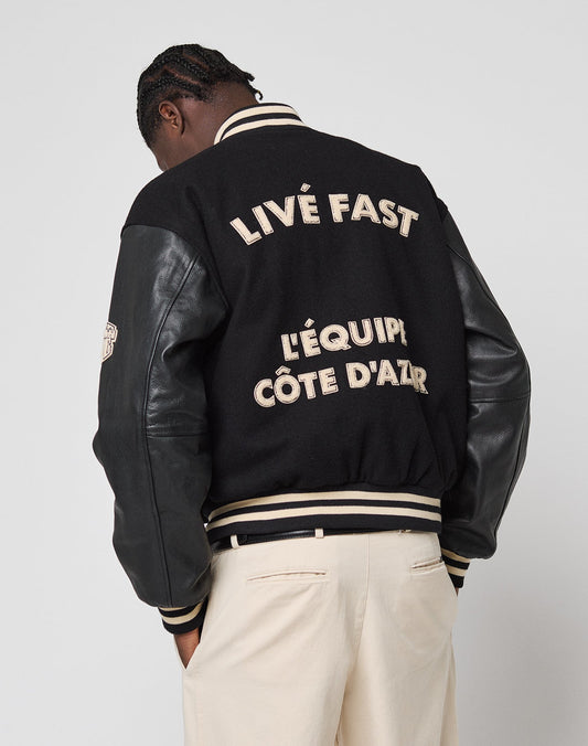 A person stands against a light background wearing cream pants and the LFDY x Schott NYC Varsity Jacket by LIVE FAST DIE YOUNG, featuring beige LIVE FAST and LÉQUIPE CÔTE DAZUR lettering on the black back.