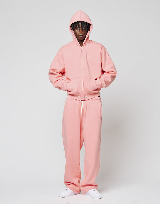 Against a plain white background, a person wears the LIVE FAST DIE YOUNG Basic Hooded Zip in light pink with matching sweatpants, hood up and zipped, paired with crisp white sneakers.