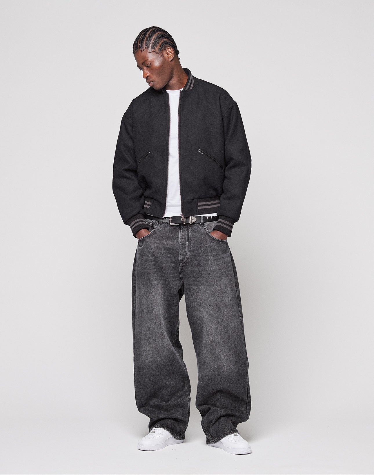 A person stands against a plain background wearing the LIVE FAST DIE YOUNG Live Fast College Jacket with Nieten Patch Logo and Metal Reißverschluss, a white t-shirt, oversized faded black Regular Fit jeans, and white sneakers, looking down with hands in pockets.