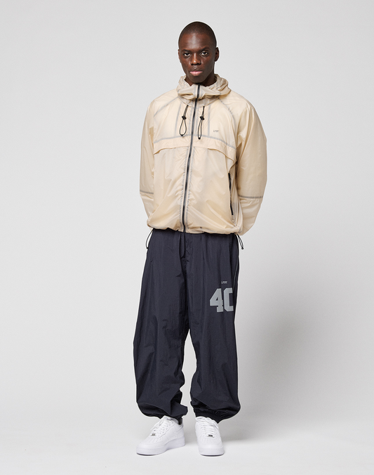 A person stands against a plain background wearing the LIVE FAST DIE YOUNG Live Fast Nylon Jacket in light beige with transparent outer fabric, loose dark navy pants with a large reflective "40" print on the left leg, and white sneakers. Hands are behind their back.