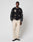 A man stands against a white background wearing a black varsity jacket with cream details, LIVE FAST DIE YOUNG Wide Chino trousers in 100% cotton, and black-and-white dress shoes. He faces forward neutrally, hands at his sides.