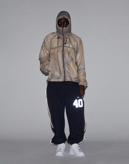 A person in a beige hooded jacket with zippered face cover, white sneakers, and black LIVE FAST DIE YOUNG LFDY Performance Trackpants featuring a reflective “40” print on the left leg stands against a plain gray background.