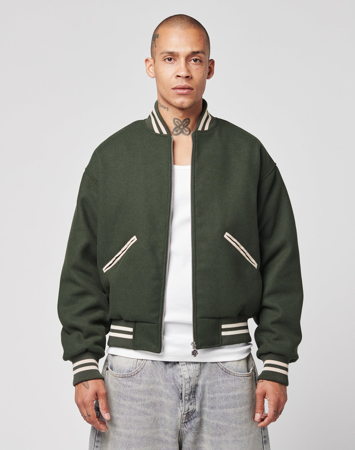 A person with a shaved head and neck tattoos wears the LIVE FAST DIE YOUNG Live Fast College Jacket in dark green with beige trim and a DY zipper, paired with a white shirt and light-wash jeans, standing against a plain light background.