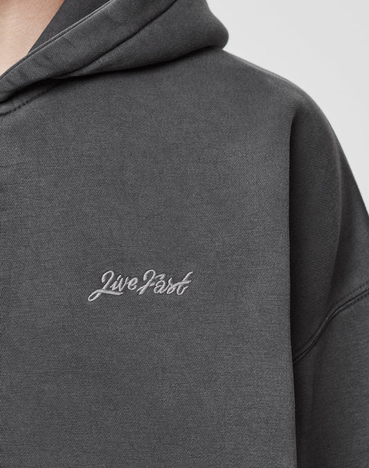 A close-up of the dark gray LIVE FAST DIE YOUNG Basic Script Hooded Zip aus Baumwolle mit Vintage-Waschung, showing the white Live Fast cursive logo on the chest. Only the upper part of the hoodie and part of the neck are visible.
