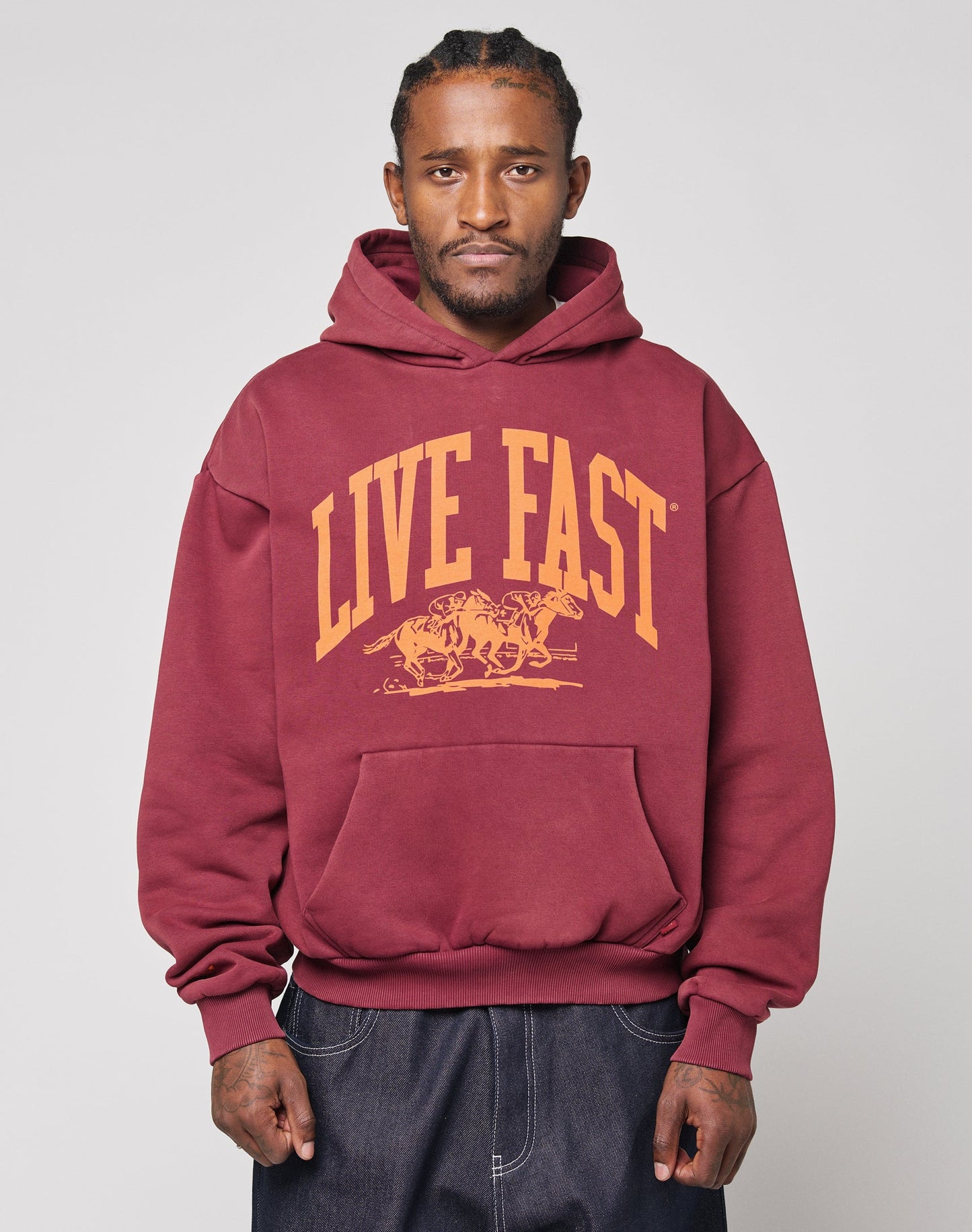 A person wears the LIVE FAST DIE YOUNG Live Fast Stallion Hooded, an oversize maroon hoodie with yellow LIVE FAST and running horses on the front, made from soft cotton and polyester, standing against a light gray background. | model