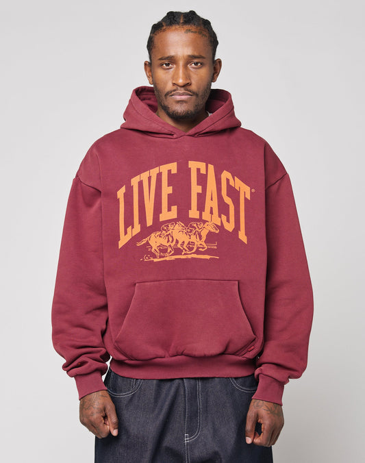 A person wears the LIVE FAST DIE YOUNG Live Fast Stallion Hooded, an oversize maroon hoodie with yellow LIVE FAST and running horses on the front, made from soft cotton and polyester, standing against a light gray background. | model
