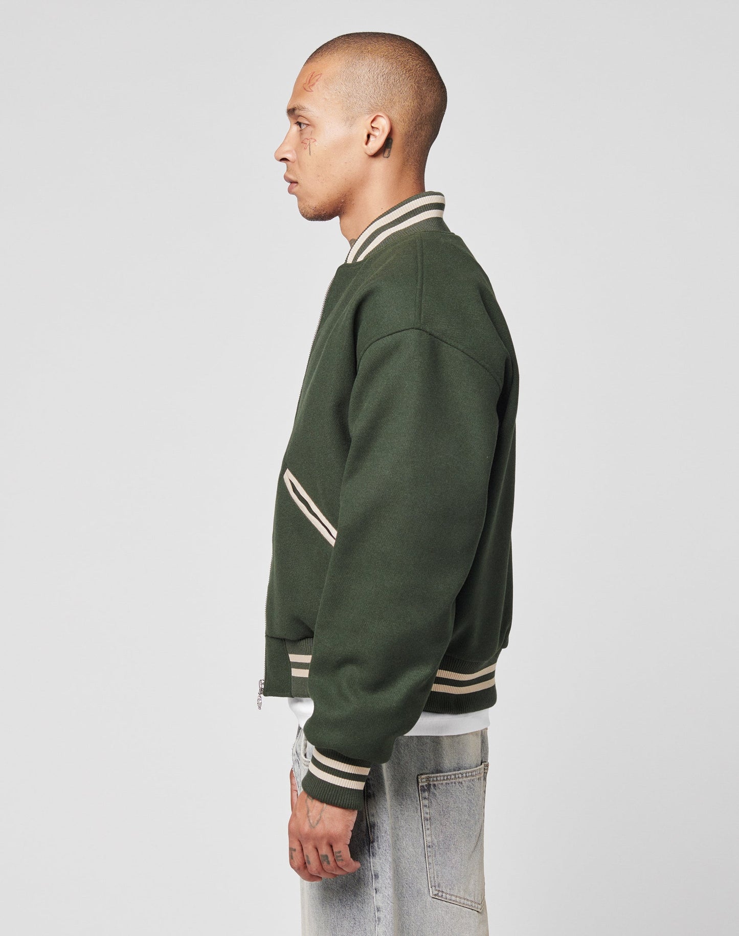 A person with a shaved head stands in profile, wearing the LIVE FAST DIE YOUNG Live Fast College Jacket in forest green with beige trim, paired with light blue jeans, against a plain light gray background.