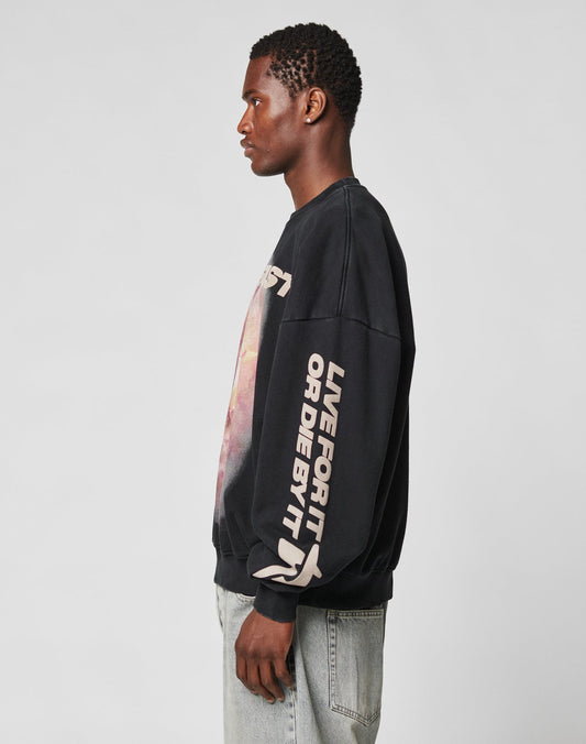 A person in profile wears the LFDY Angel Sweater by LIVE FAST DIE YOUNG—an oversized black sweatshirt with LIVE FOR IT OR DIE BY IT in bold white sleeve print—paired with light blue jeans, set against a plain gray background.