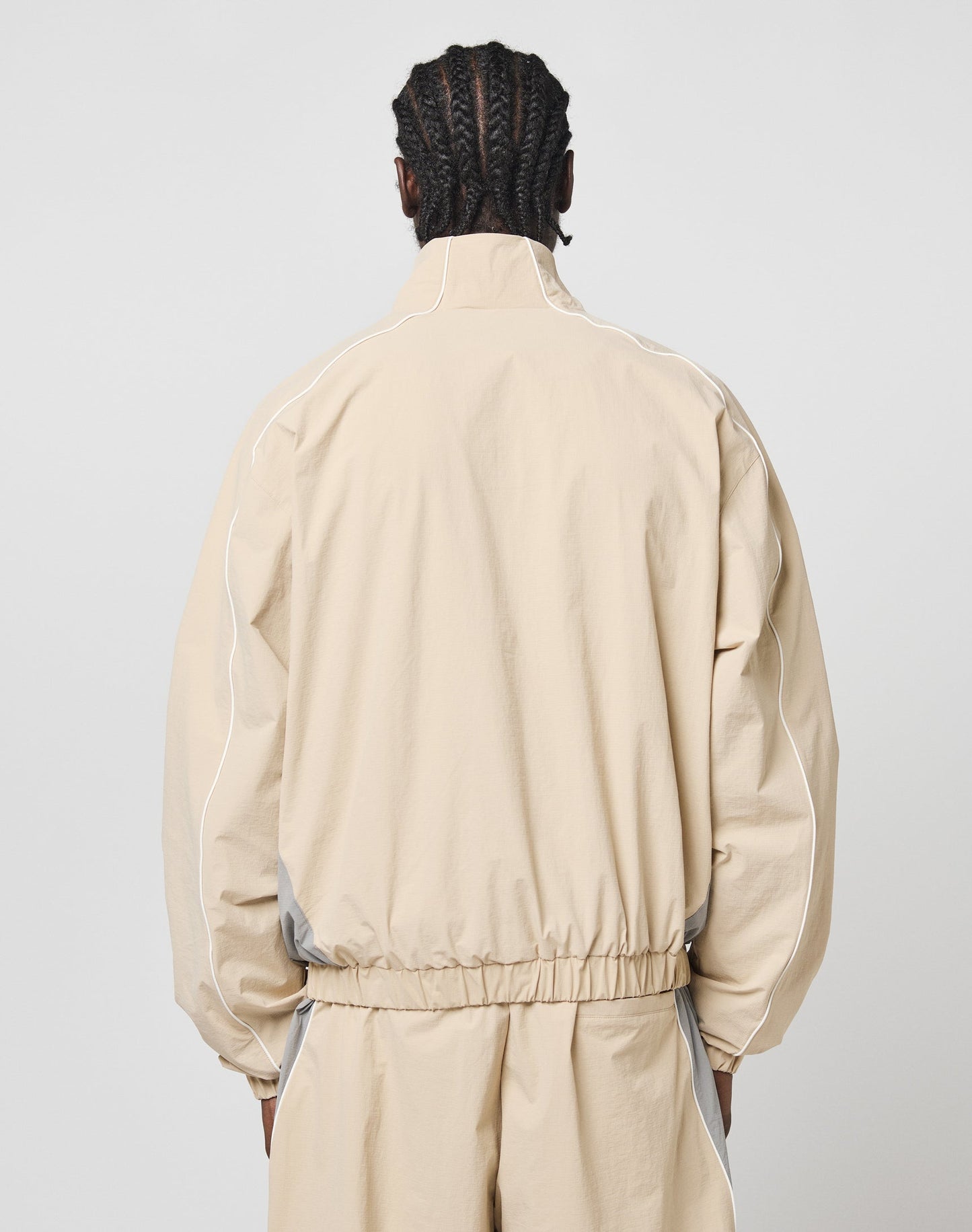 A person with braided hair, wearing the LIVE FAST DIE YOUNG Basic Nylon Trackjacket in beige with white piping and a regular fit, stands against a plain light gray background facing away from the camera.