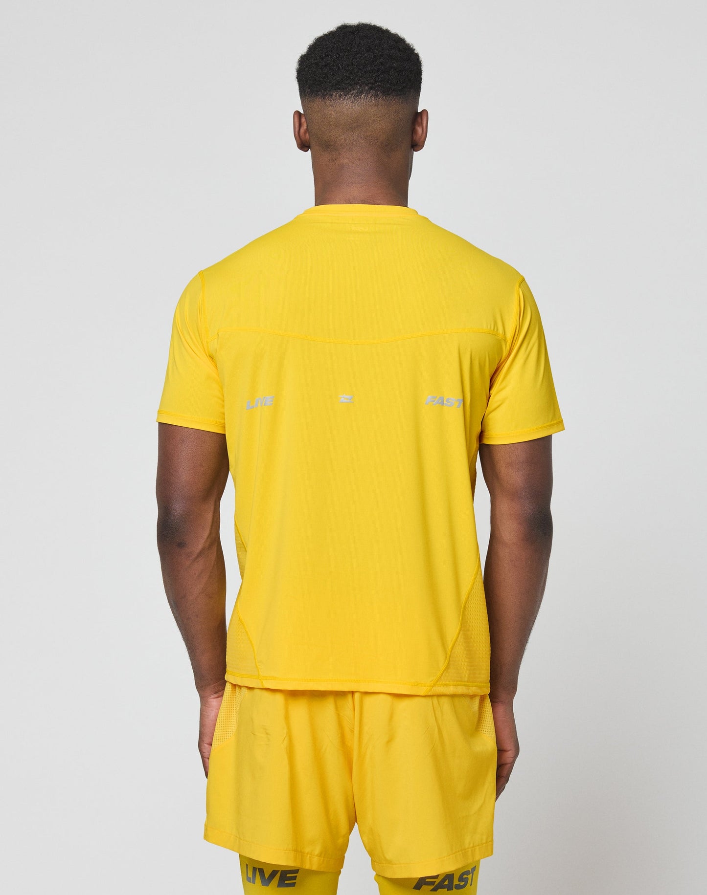 A man faces away from the camera in a bright yellow Live Fast Performance Tee by LIVE FAST DIE YOUNG. The light gray backdrop highlights its slim fit nylon-spandex fabric and reflective print on the upper back.