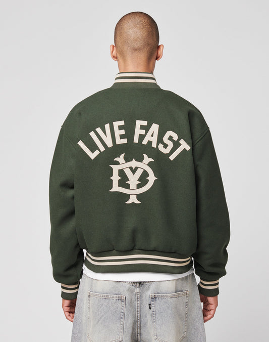 A person with a shaved head is seen from behind wearing the Live Fast College Jacket by LIVE FAST DIE YOUNG. The dark green varsity features white trim and large LIVE FAST text above a LY logo embroidered on the back. | model