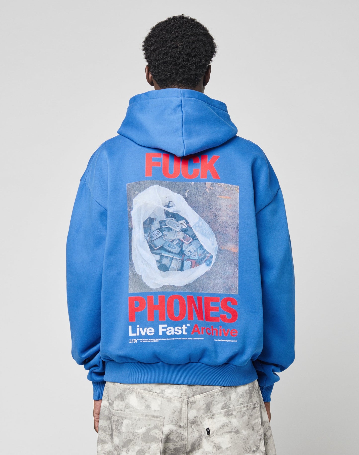 A person faces away in an oversized LFDY F*ck Phones Hoodie by LIVE FAST DIE YOUNG, featuring bold red “FUCK PHONES” text and a graphic of a plastic bag with old phones above “Live Fast Archive,” crafted from soft cotton.