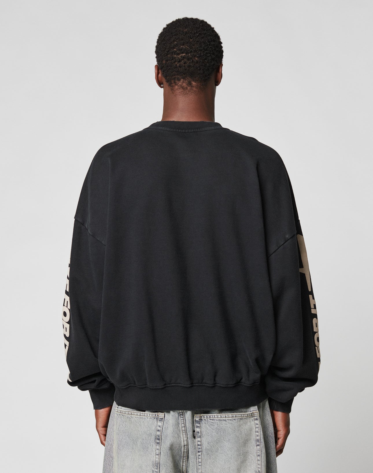 A person with short hair, wearing the LFDY Angel Sweater by LIVE FAST DIE YOUNG and light blue Oversize Fit jeans, stands with their back to the camera against a plain gray background.