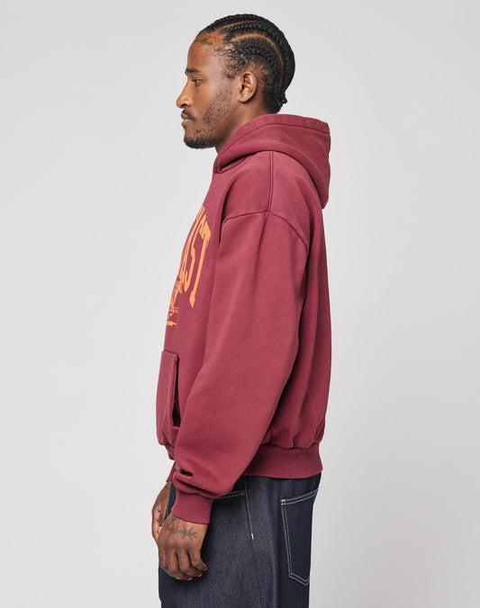 A man with cornrow braids stands in profile, wearing the LIVE FAST DIE YOUNG Live Fast Stallion Hooded in maroon with yellow writing, made from a soft cotton-polyester blend, paired with dark jeans. The background is plain light gray.