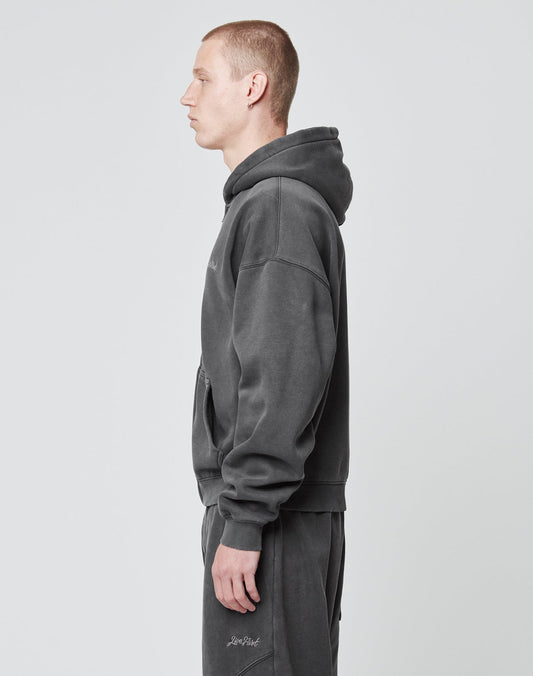 A person with short hair stands in profile, facing left, wearing the LIVE FAST DIE YOUNG Basic Script Hooded Zip—dunkelgrau aus Baumwolle mit Vintage-Waschung und dezentem Logo-Stick—vor einem schlichten hellgrauen Hintergrund.