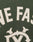 Close-up of green fabric with white stitched applique letters spelling NE FAS, ornate white design below, and a DY-Reißverschluss for contrast—details from the LIVE FAST DIE YOUNG Live Fast College Jacket.