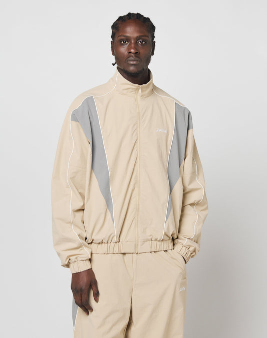 A person models the LIVE FAST DIE YOUNG Basic Nylon Trackjacket in beige with gray and white accents, standing against a plain white background and looking at the camera with a neutral expression.