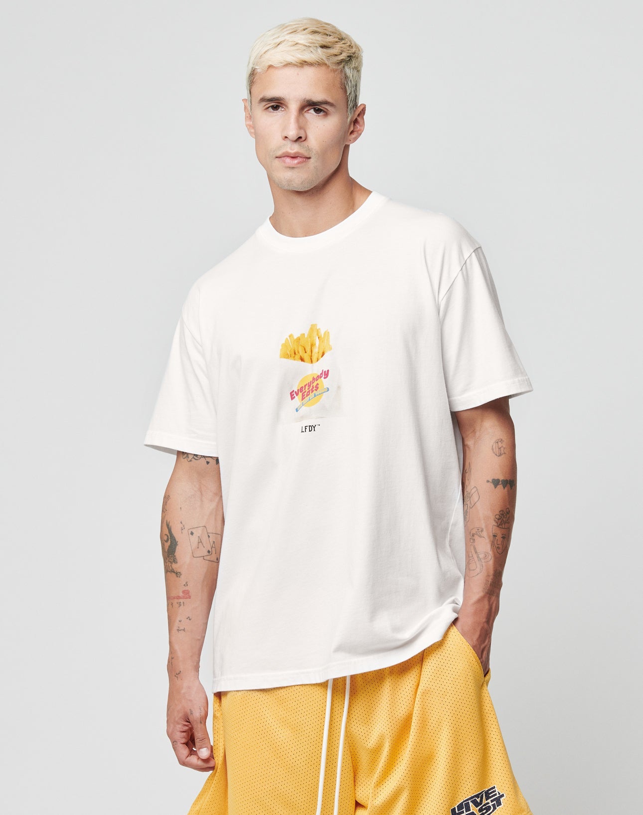 A man with short blond hair wears the LIVE FAST DIE YOUNG LFDY Fries Tee, a regular fit white t-shirt with a French fries graphic, and yellow basketball shorts. He stands against a light gray background, left hand in his pocket.