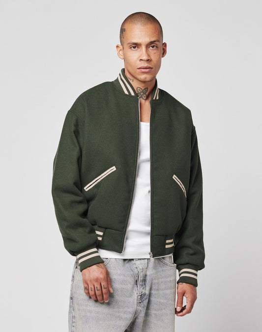 A man with a shaved head and neck tattoos wears the LIVE FAST DIE YOUNG Live Fast College Jacket, featuring contrasting stripes and back embroidery, over a white tank top and light blue jeans against a plain gray background.
