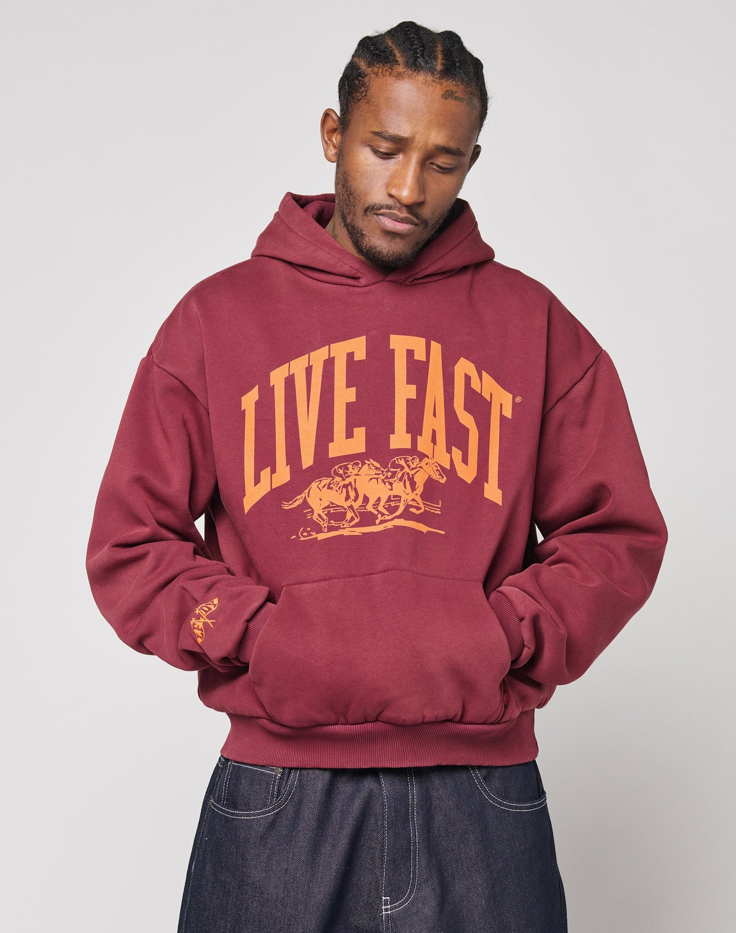Someone wearing the Live Fast Stallion Hooded by LIVE FAST DIE YOUNG, a maroon oversized hoodie featuring yellow LIVE FAST text and two tigers graphic, made from a soft cotton-polyester blend, stands with hands in the pocket and looks down. | plain