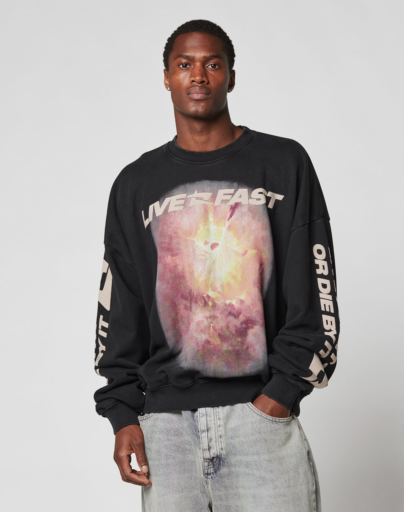 A man wears the LFDY Angel Sweater by LIVE FAST DIE YOUNG, an oversized black cotton sweatshirt with LIVE FAST text, a colorful abstract graphic, and white sleeve detail. He pairs it with light-wash jeans against a gray background.