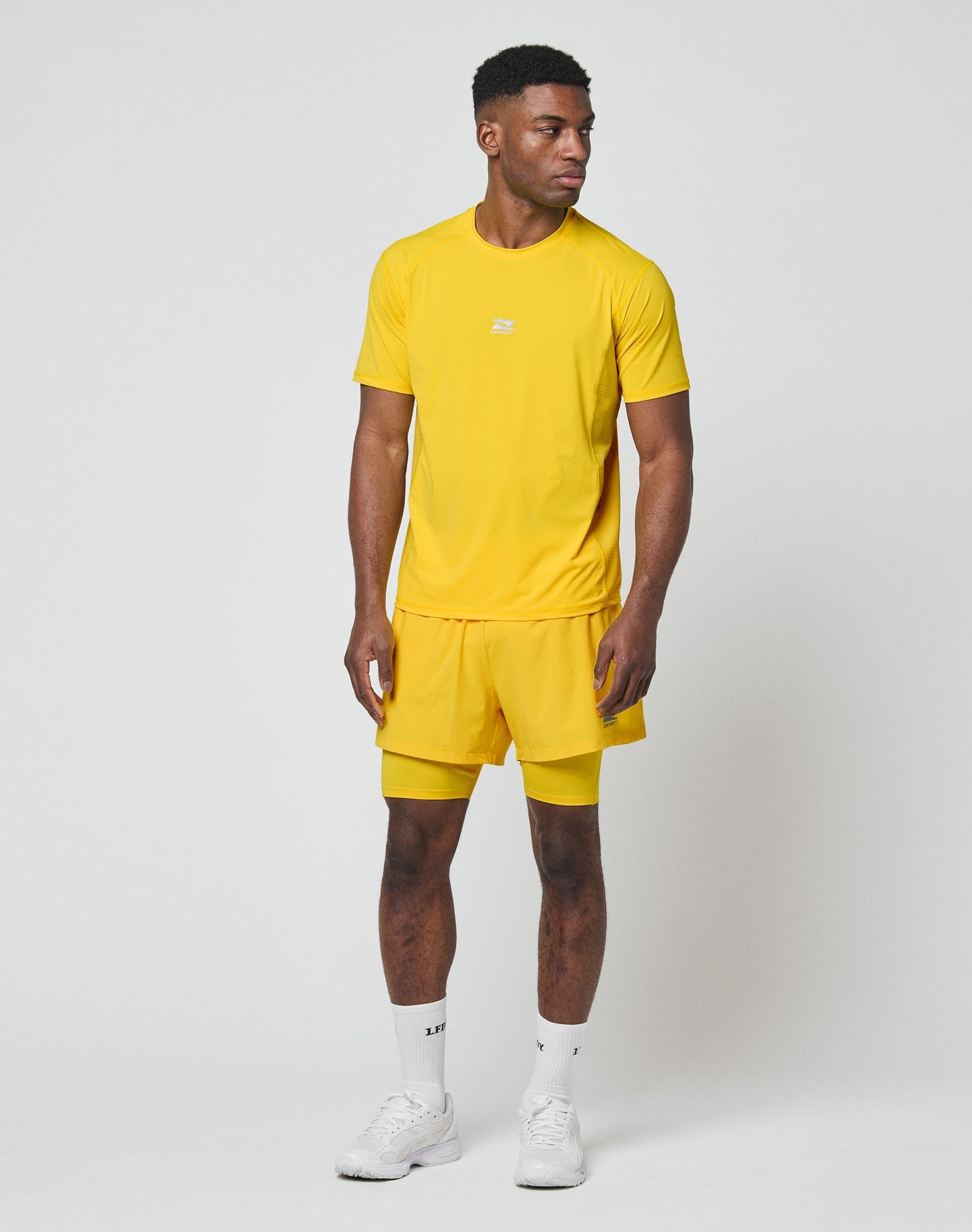 A man stands against a plain white background wearing the LIVE FAST DIE YOUNG Live Fast Performance Tee in bright yellow with matching shorts, white socks, and sneakers. He looks slightly left with his arms relaxed at his sides.