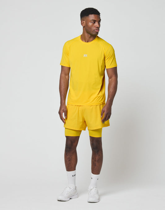 A man stands against a plain white background wearing the LIVE FAST DIE YOUNG Live Fast Performance Tee in bright yellow with matching shorts, white socks, and sneakers. He looks slightly left with his arms relaxed at his sides.