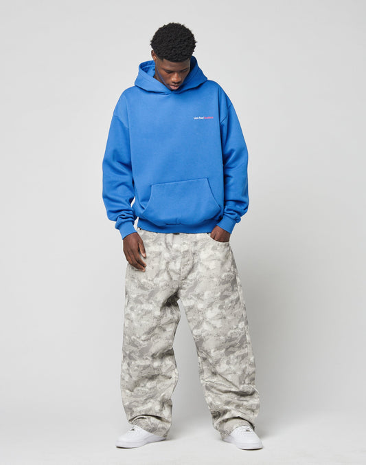 A man stands against a plain background wearing the LIVE FAST DIE YOUNG LFDY F\*ck Phones Hooded in bright blue, paired with light gray camo baggy pants and white sneakers, looking down with his hands in his pockets.