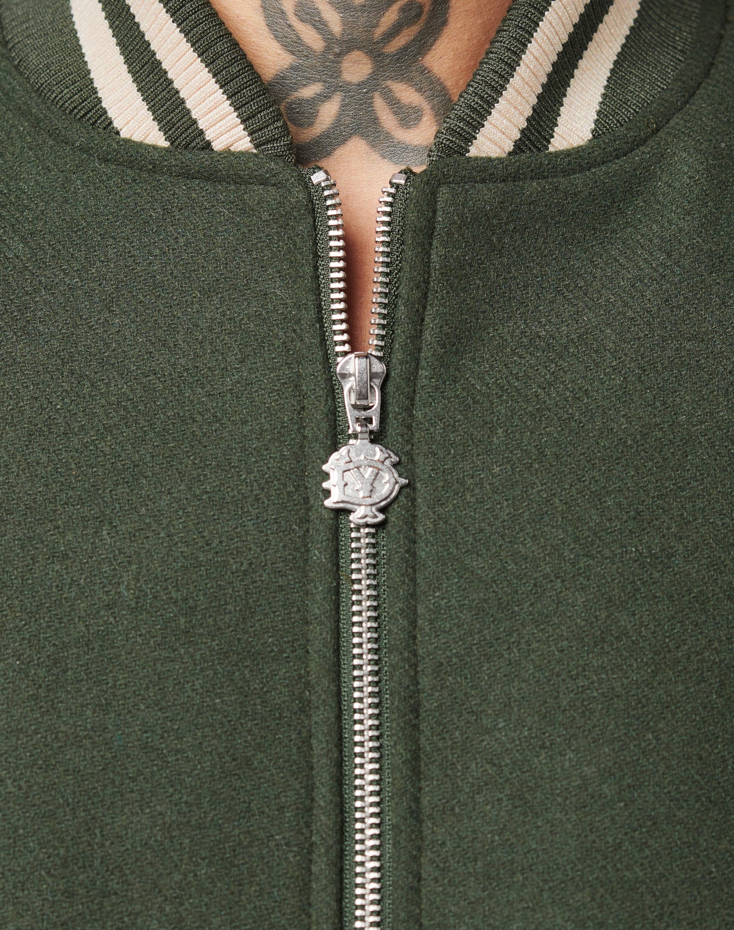 Close-up of the Live Fast College Jacket by LIVE FAST DIE YOUNG in green, featuring a DY zipper partly unzipped to show a black floral tattoo on the upper chest and neck. The white-striped ribbed collar adds contrasting color for visual appeal.