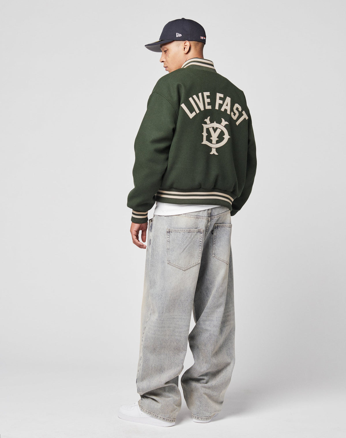 A person wears the LIVE FAST DIE YOUNG Live Fast College Jacket with Rückenstick and a circular back logo, loose light-wash jeans, white sneakers, and a dark baseball cap against a plain light background.