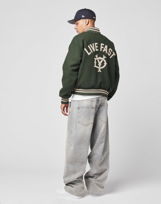 A person wears the LIVE FAST DIE YOUNG Live Fast College Jacket with Rückenstick and a circular back logo, loose light-wash jeans, white sneakers, and a dark baseball cap against a plain light background.
