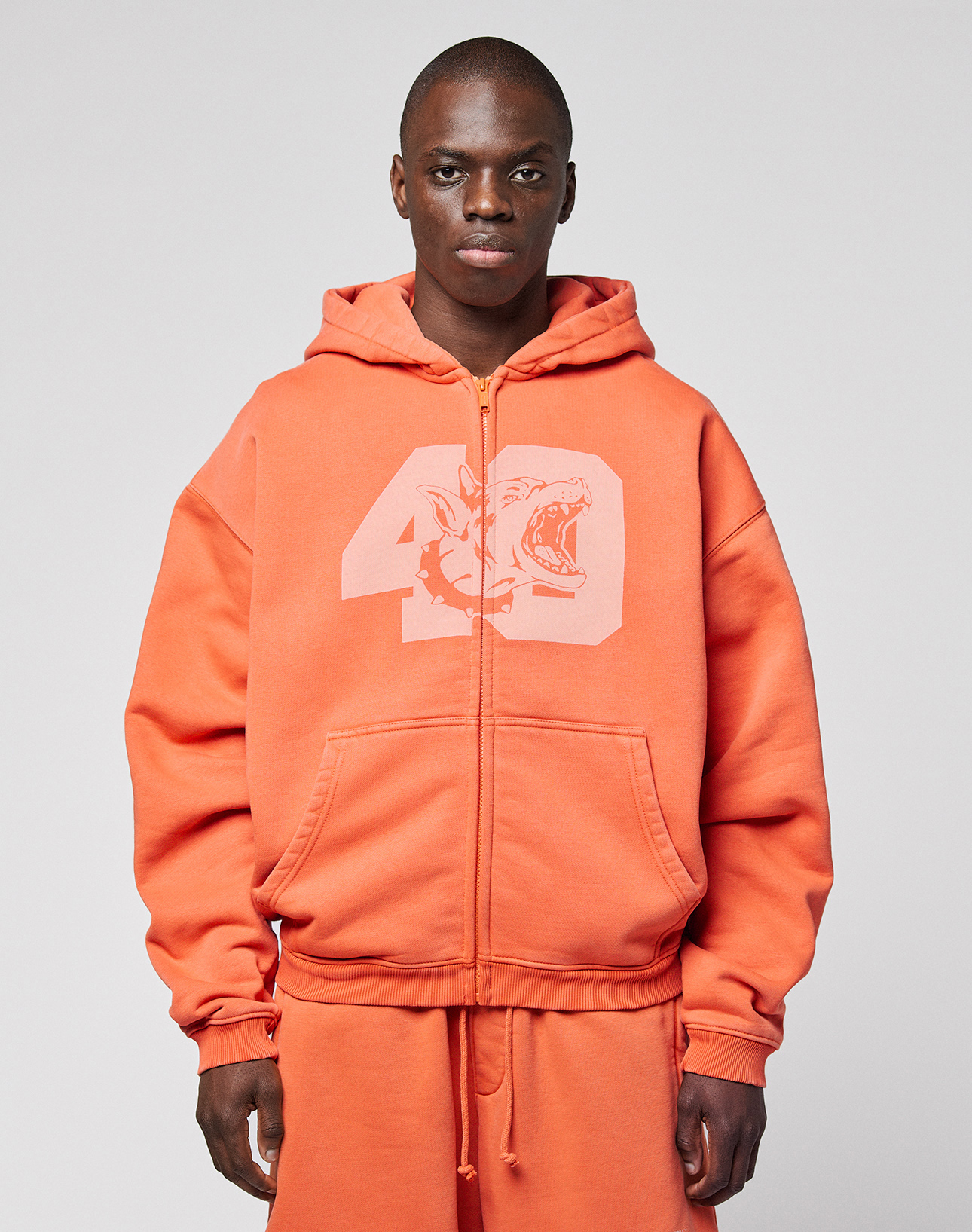 A person wearing the orange LFDY 40 Hooded Zip by LIVE FAST DIE YOUNG, featuring a large number 49 and shark front print, stands against a plain light gray background. | model