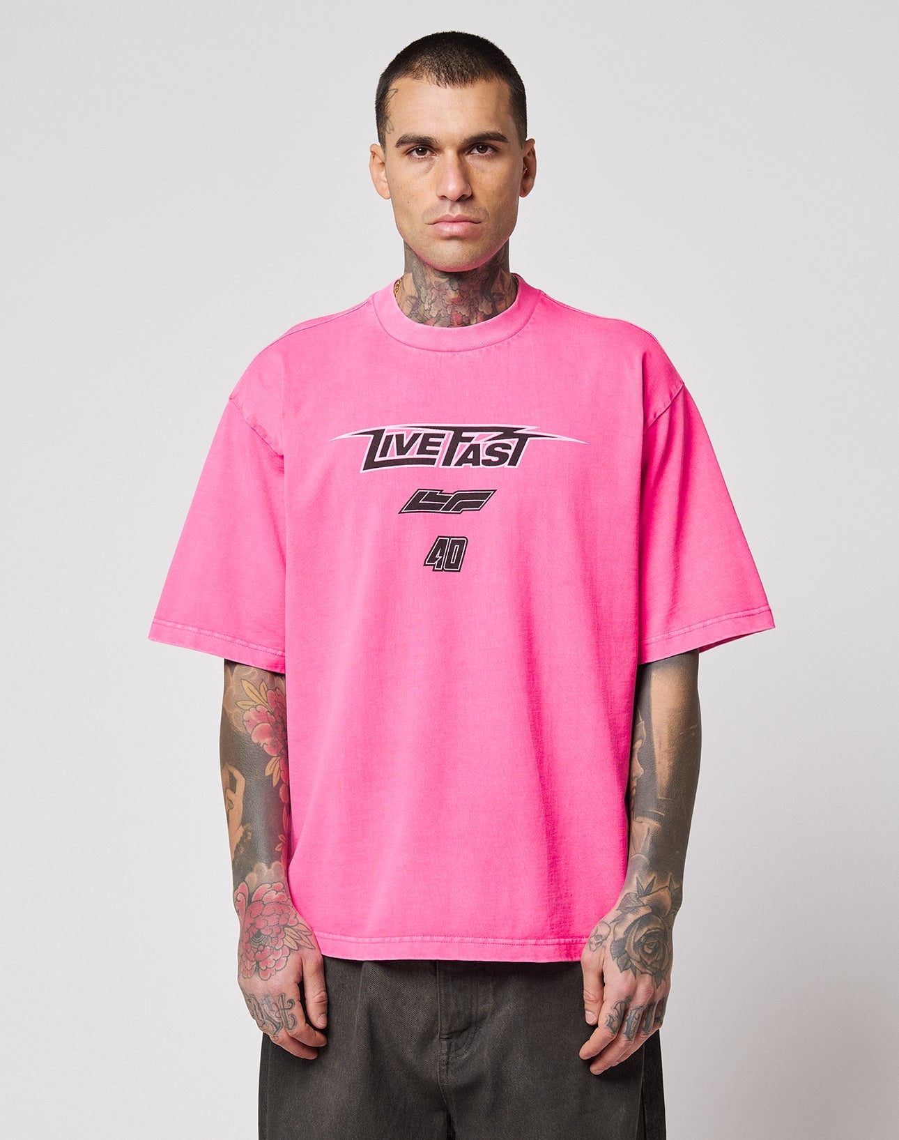 A man with tattoos wears the LFDY Triple Racing Tee by LIVE FAST DIE YOUNG—an oversize, bright pink 100% cotton T-shirt with a bold multilogo LIVE FAST front print—standing against a plain light gray background.