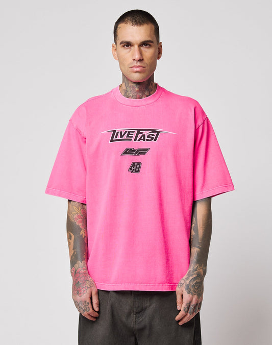 A man with tattoos wears the LFDY Triple Racing Tee by LIVE FAST DIE YOUNG—an oversize, bright pink 100% cotton T-shirt with a bold multilogo LIVE FAST front print—standing against a plain light gray background. | model