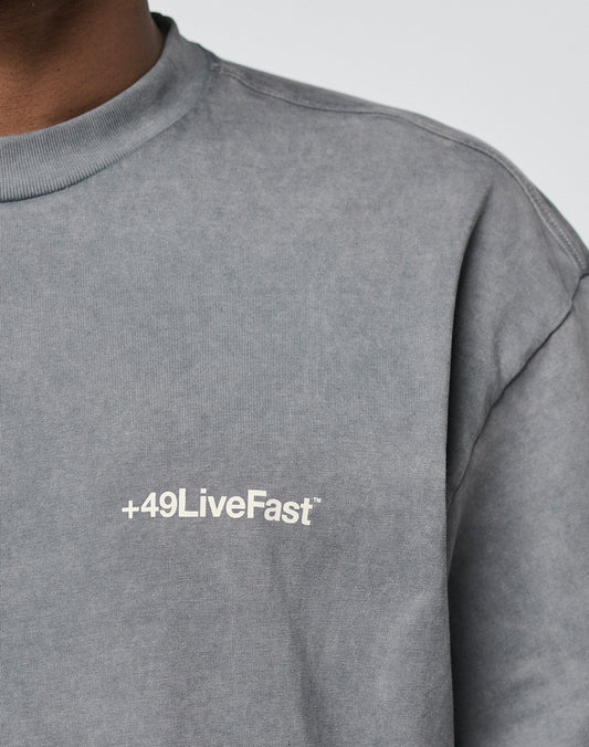 A person wears the LIVE FAST DIE YOUNG Basic +49 Chest Tee—an oversize gray shirt made from 100% Baumwolle, featuring a white +49LiveFast logo on the left chest. Only part of the upper body and shirt text are visible.