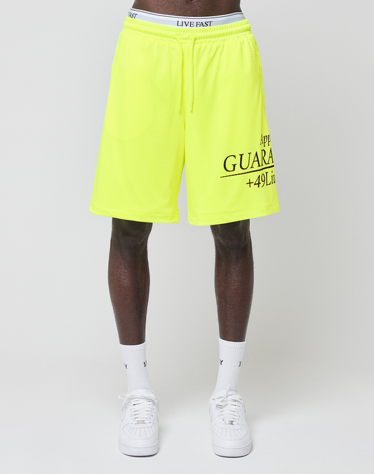 A person wears bright yellow LFDY Guaranteed Mesh Shorts by LIVE FAST DIE YOUNG with an elastic waistband, black text, white socks, and white sneakers, standing against a plain light background.