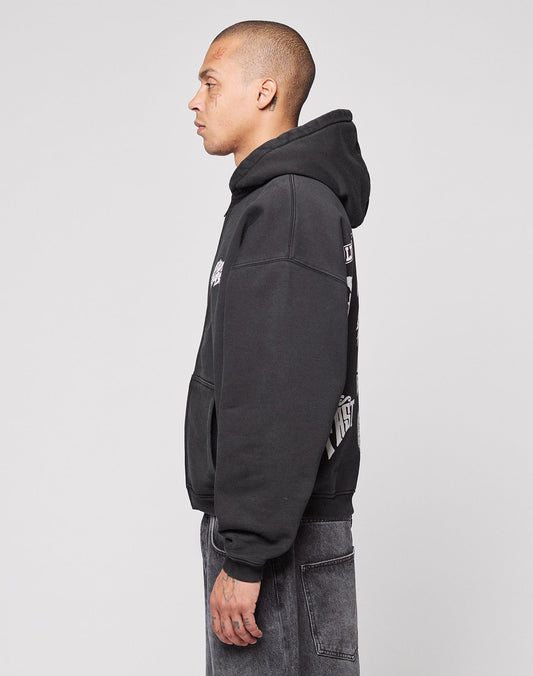A person with short hair stands in profile wearing a black LIVE FAST DIE YOUNG LFDY Logo Hooded Zip in Oversize Fit, featuring graphic designs on the back and sleeve, paired with faded black jeans against a light gray background.
