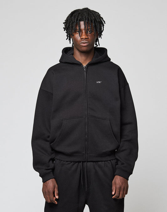 A person with medium-dark skin and short, twisted hair wears the LIVE FAST DIE YOUNG Basic Hooded Zip in black with matching pants, standing against a plain light gray background. | model