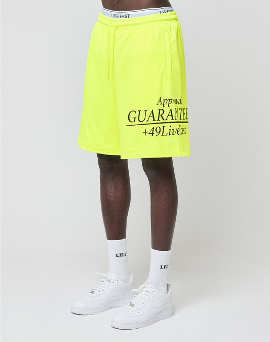 A person wearing neon yellow LFDY Guaranteed Mesh Shorts by LIVE FAST DIE YOUNG, white socks with LFDT text, and white Nike sneakers stands against a plain light background. Only the lower body is shown. | model
