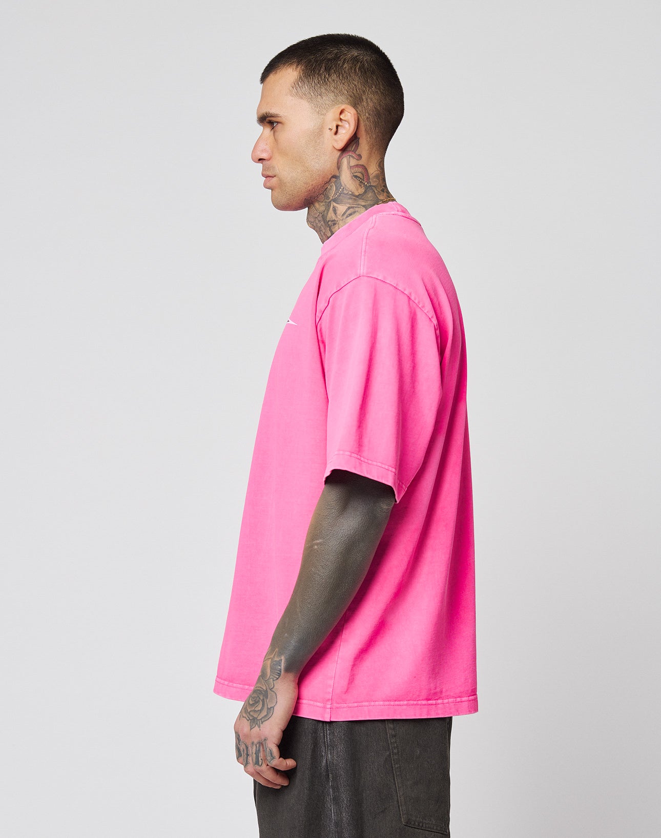 A person with tattoos on their neck and arms stands sideways, wearing a bright pink LFDY Triple Racing Tee by LIVE FAST DIE YOUNG and dark shorts against a plain light gray background.