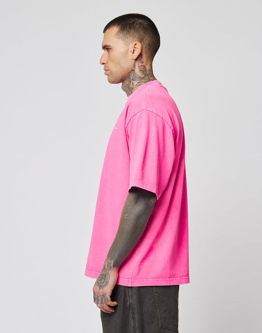 A person with tattoos on their neck and arms stands sideways, wearing a bright pink LFDY Triple Racing Tee by LIVE FAST DIE YOUNG and dark shorts against a plain light gray background.