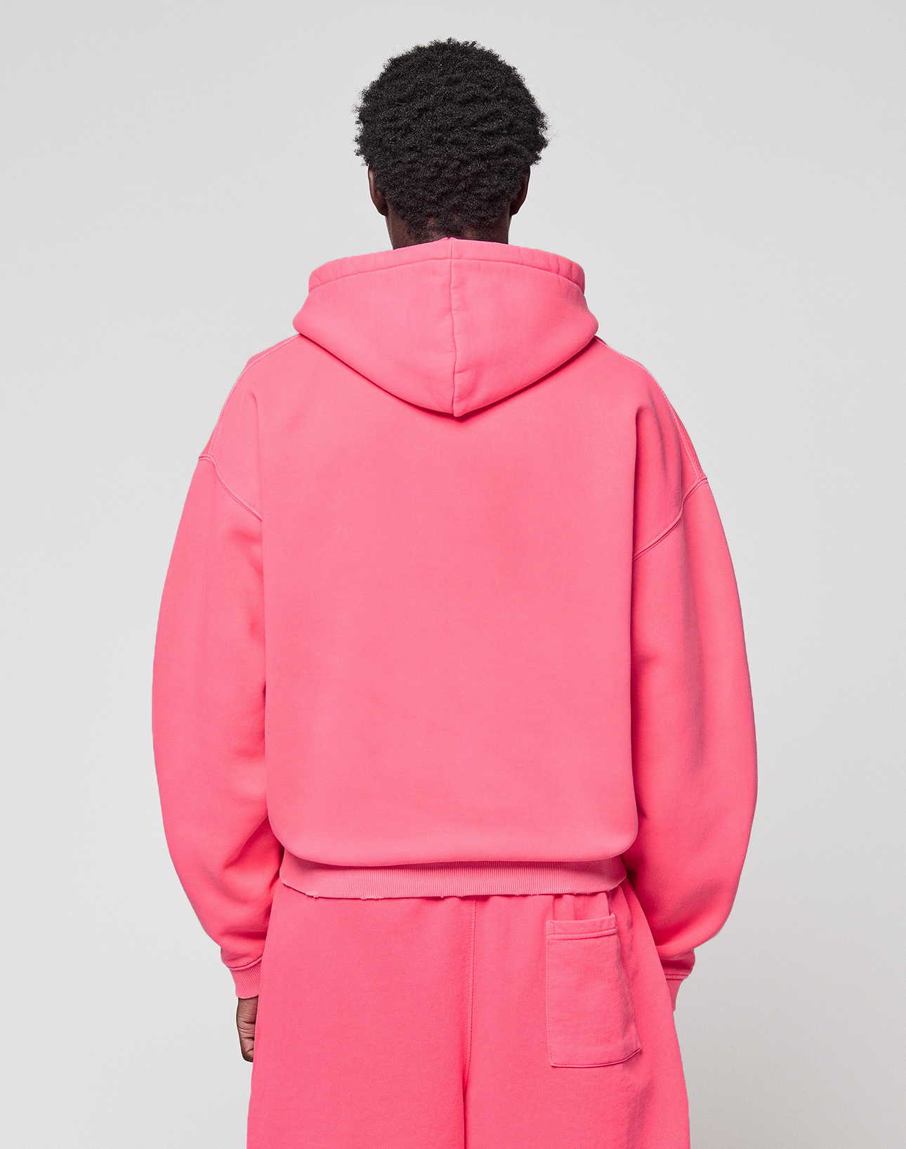 A person faces away from the camera, wearing a bright pink LIVE FAST DIE YOUNG Basic Hooded Zip oversize hoodie and matching sweatpants with a pocket, set against a plain light background.
