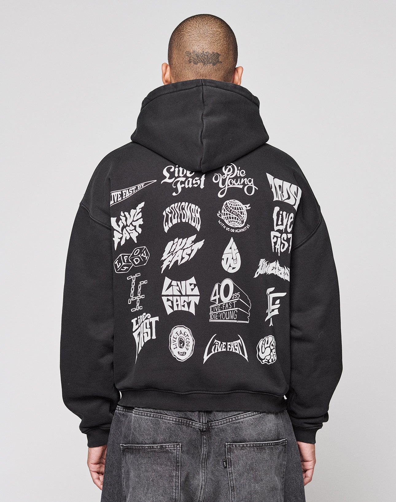 A person faces away from the camera in a black LIVE FAST DIE YOUNG LFDY Logo Hooded Zip, an oversized Baumwolle hoodie with multilogo white graphics and “Live Fast” and “Die Young” phrases on the back, set against a plain gray background.
